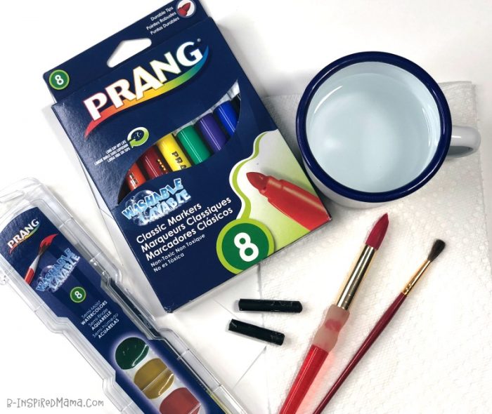 A photo of the supplies needed for a watercolor butterfly painting, including Prang markers, watercolor paint set, a paintbrush and cup of water, and black crayons.
