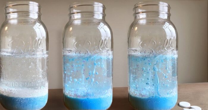 A photo of 3 glass mason jars filled with a fizzing and bubbling light blue, sparkly water-and-oil mixture that looks like a snowstorm. A couple small white Alka Seltzer Effervescent Tablet sit on the table next to the jars.