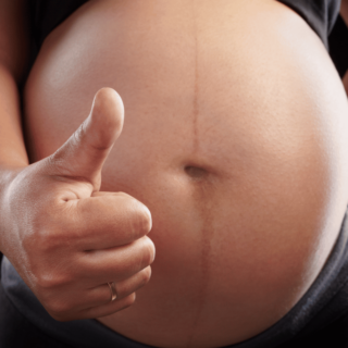 A photo of a pregnant person's belly with their hand extended in front of it giving a thumbs up.