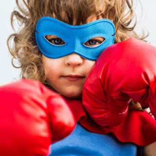A photo of a determined young girl in a blue superhero mask holding red boxing gloves up to her face.