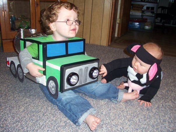 A young boy is sitting on the floor, wearing a homemade milk tanker truck Halloween costume. A toddler wearing a homemade cow costume sits nearby and points to the working headlights on the milk truck costume.
