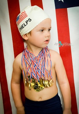 A young boy stands in front of an American flag wearing an easy DIY Halloween costume consisting of dark swim trunks, a swim cap with a flag on it, and multiple pretend gold medals made out of red, white, and blue striped ribbon and gold-painted cardboard.