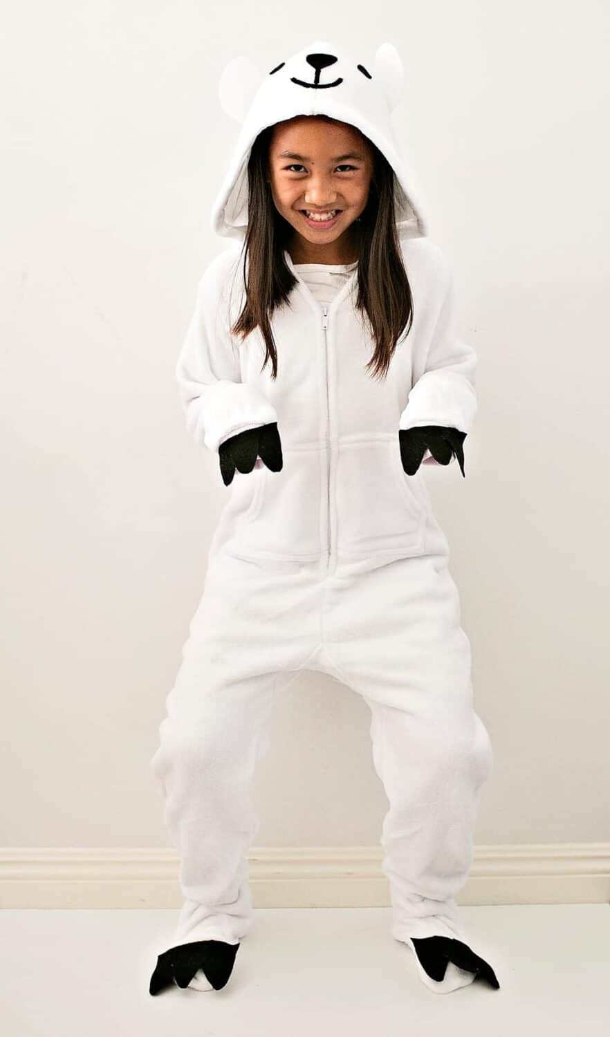 A smiling girl stands wearing a homemade polar bear Halloween costume consisting of white onesie pajamas with black felt paw details attached to each foot and each hand along with white felt ears and black felt nose and eyes attached to the hood.