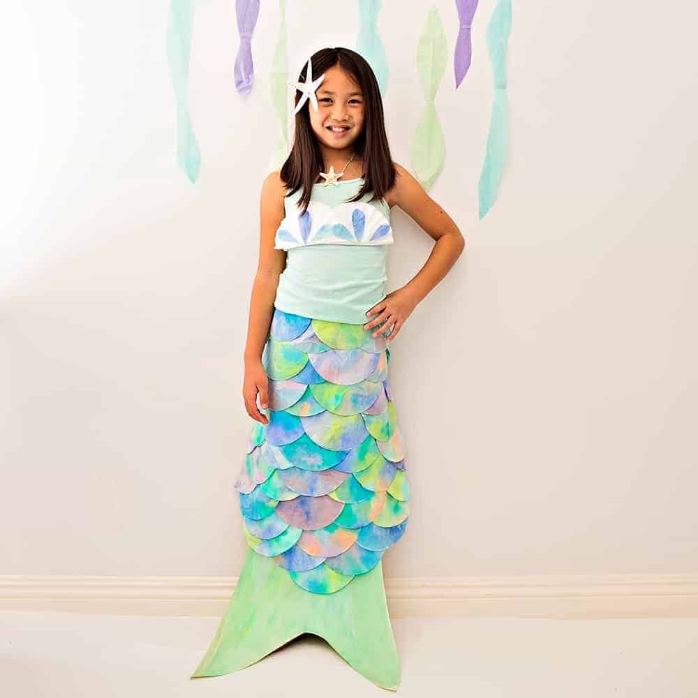 A smiling girl stands wearing a DIY mermaid creative Halloween costume consisting of a mint green and teal mermaid tail made out of cardboard and colorful coffee filters.