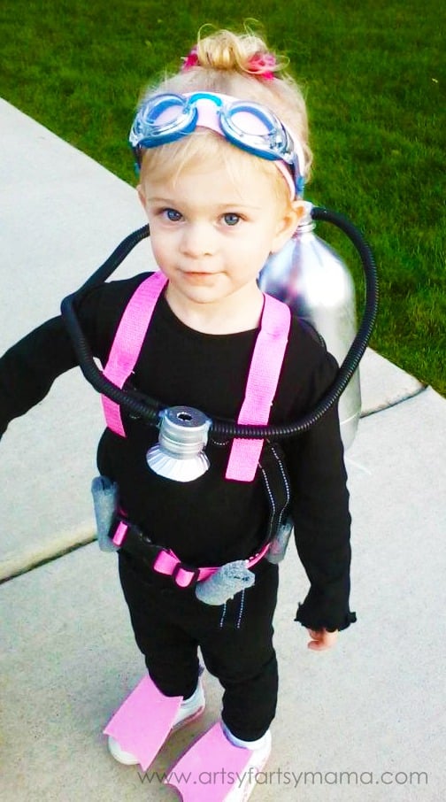 A little girl stands outside wearing a DIY scuba diver Halloween costume consisting of goggles on her head and black clothing with pink felt straps and flippers on her shoes along with a DIY air tank made out of recycled plastic soda bottles painted silver.
