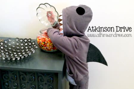 A child stands wearing a DIY Halloween costume made to look like a shark. The costume consists of a gray hooded sweatshirt with a black shark fin coming off the back, black felt eyes on the hood, and white felt teeth around the opening of the hood.