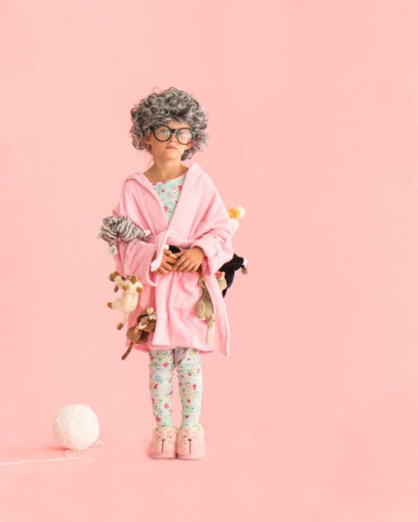 A young girl stands wearing a fun creative Halloween costume including pajamas, slippers, a pink robe with multiple plush toy cats attached to it, a gray old lady wig, and dark round eyeglasses.
