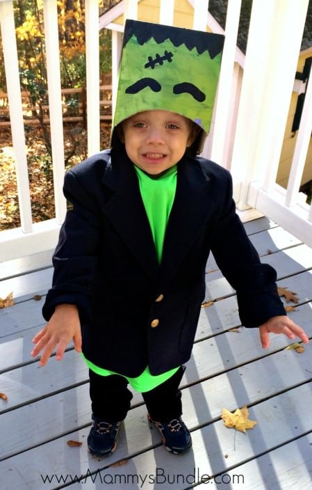 A smiling child stands outside on a porch wearing a DIY Halloween costume to look like Frankenstein, including a cardboard box hat painted bright green with black eyebrows, stitched, and hair. He also wears a bright green t-shirt, black pants, and an oversized black suit jacket.