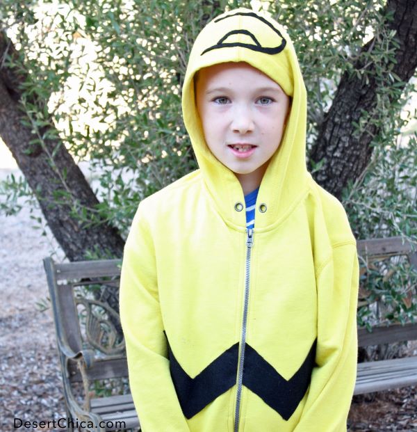 A happy child stands outside wearing a DIY Charlie Brown Halloween costume consisting of a yellow hooded sweatshirt with a black zigzag line across the waist and a black line hair accent on the top of the hood.