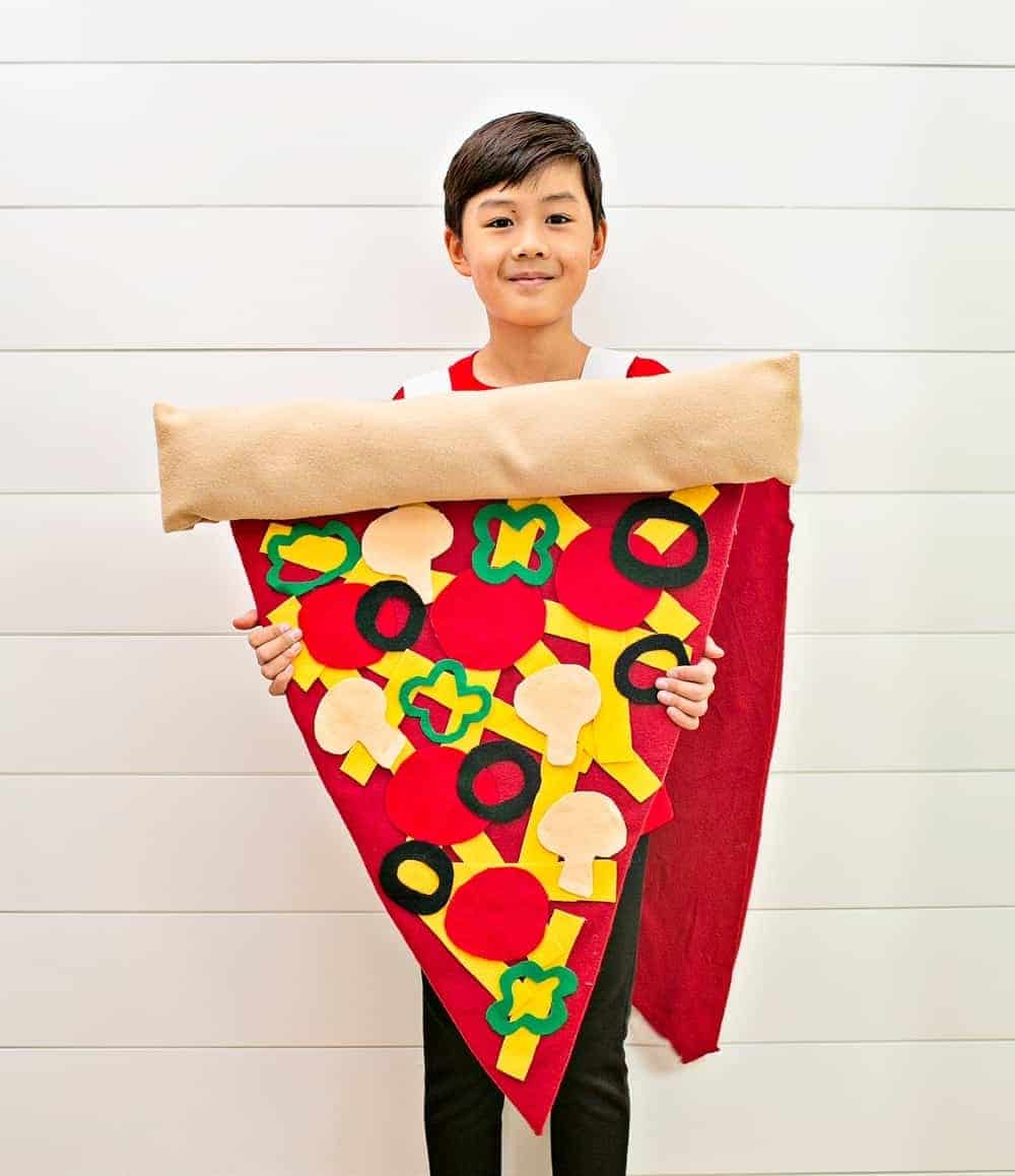 A child stands wearing a fun creative Halloween costume made out of cardboard and felt to look like a slice of pizza with various toppings including cheese, mushrooms, olives, green peppers, and pepperoni.