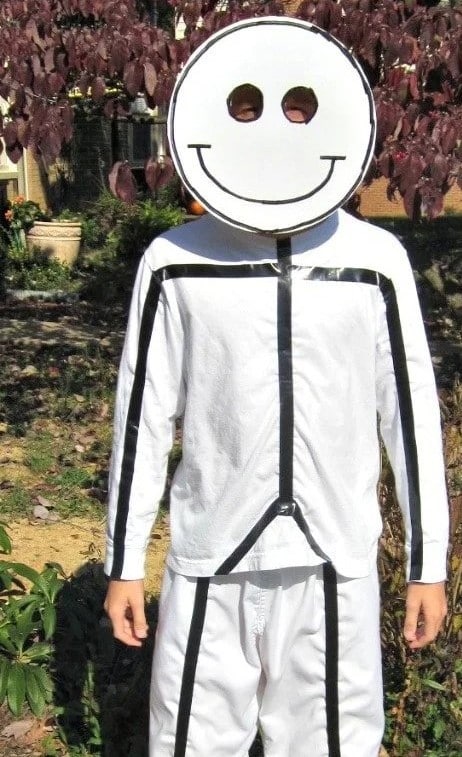 A child stands outside wearing a simple but creative Halloween costume consisting of white clothing with black tape on it in the shape of a stick figure along with a round white cardboard mask with black lines on it in the form of a smiley face.