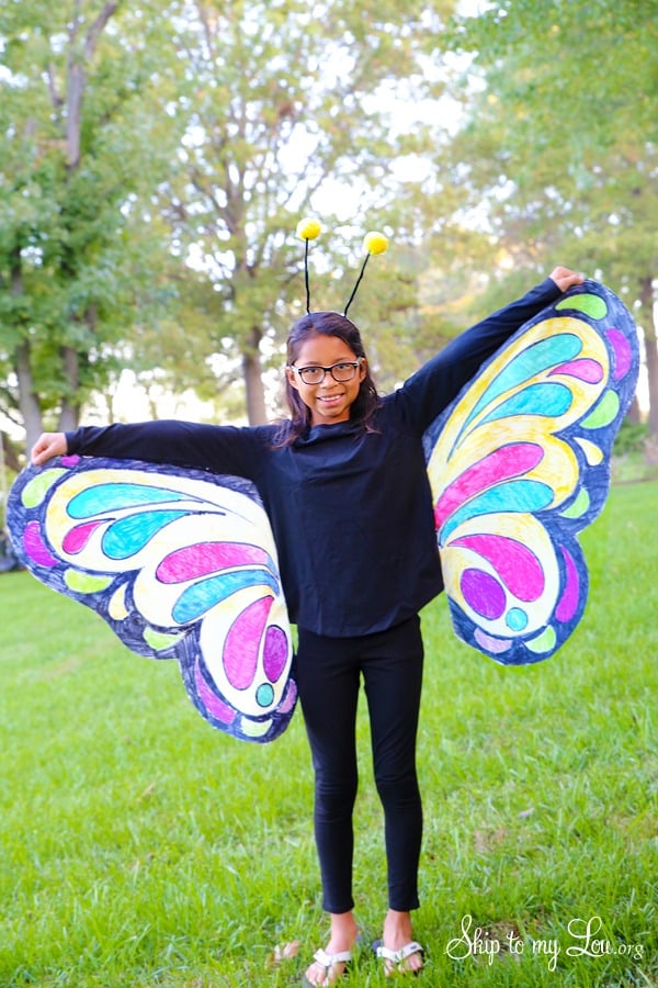 A smiling girl stands outside wearing a creative Halloween costume, a butterfly with homemade colorful fabric butterfly wings, black pants and shirt, and a simple homemade antenna headband.