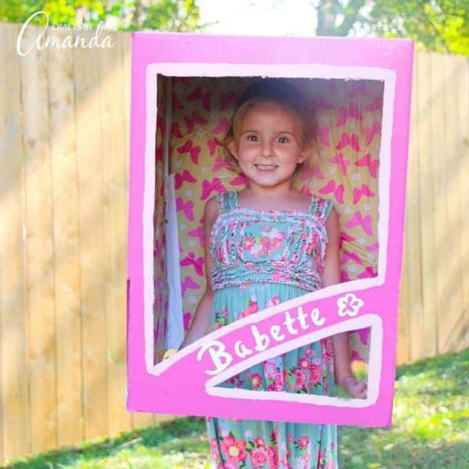 A young smiling girls stands outside wearing a DIY Barbie Doll in a Box Halloween costume which consists of a large pink box cut and painted to look like a Barbie box.