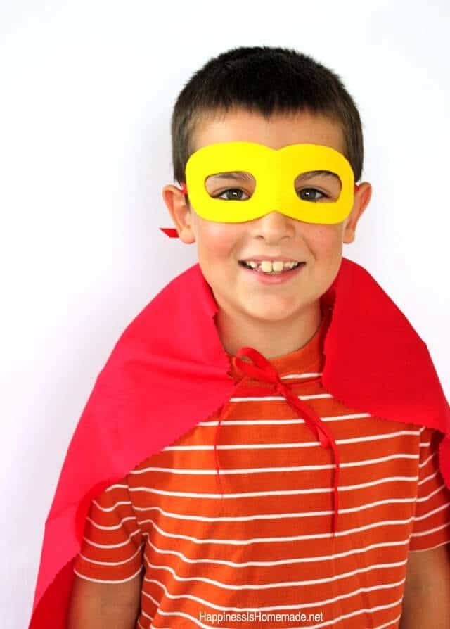 A child stands smiling, wearing a simple superhero Halloween costume consisting of a red striped shirt, a DIY red felt cape, and homemade yellow felt eye mask.
