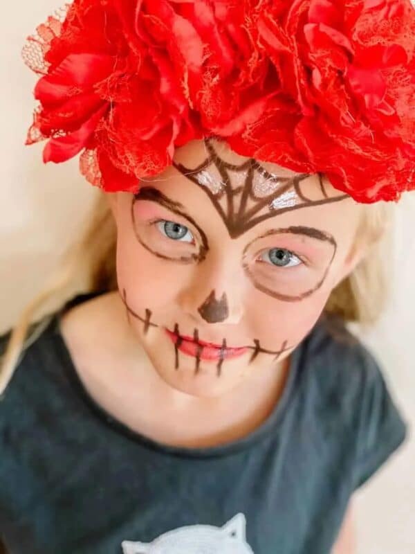 A photo of a smiling girl in a DIY sugar skull costume with her face painted to look like a sugar skull, wearing a red floral headband.