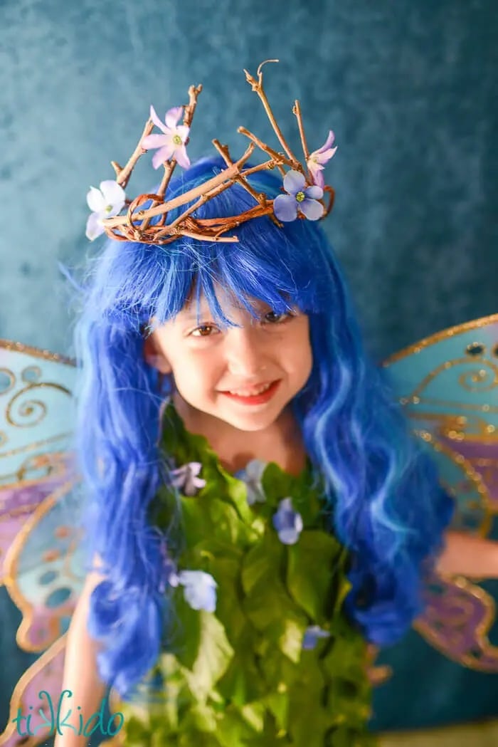 A smiling child stands wearing a homemade fairy Halloween costume consisting of fairy wings, a dress covered in fake flowers and leaves, a long blue-haired wig, and a twig and flower crown.
