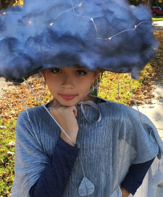 A child stands outside wearing a homemade rain cloud Halloween costume consisting of a simple silver shawl and a hat made out of white and gray fiber fill, fairy lights, and hanging silver raindrops.
