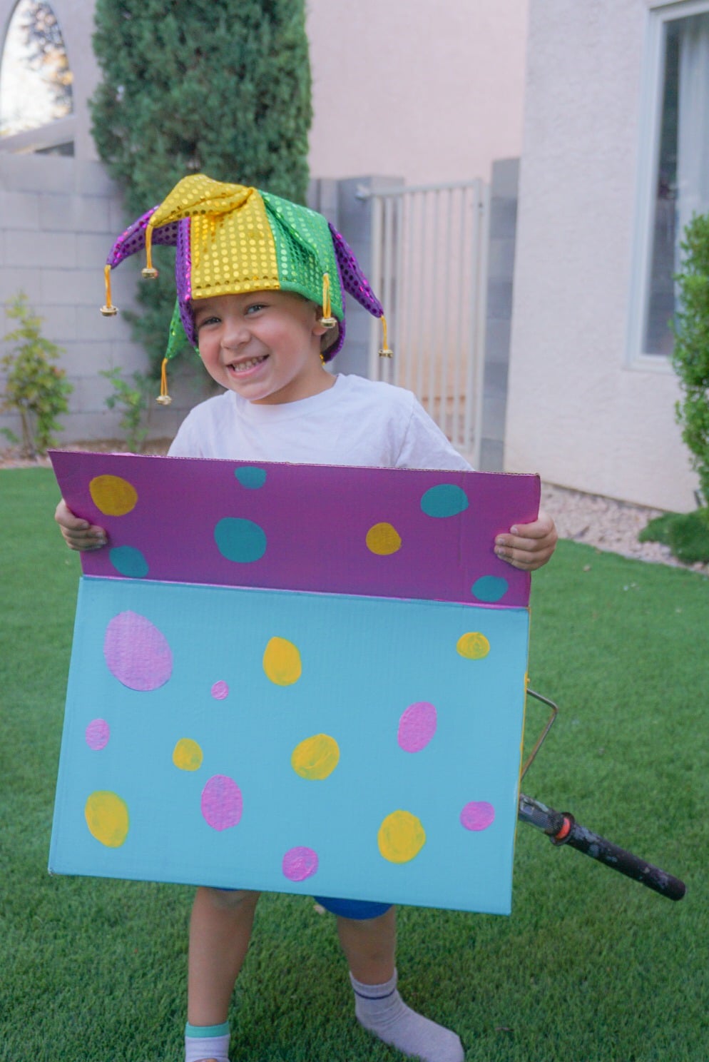 A smiling child stands outside wearing a Jack-in-the-Box creative Halloween costume made out of a cardboard box painted with colorful blue, purple, and yellow polka dots. The child also wears a white t-shirt and a whimsical purple, yellow, and green jester-style hat.