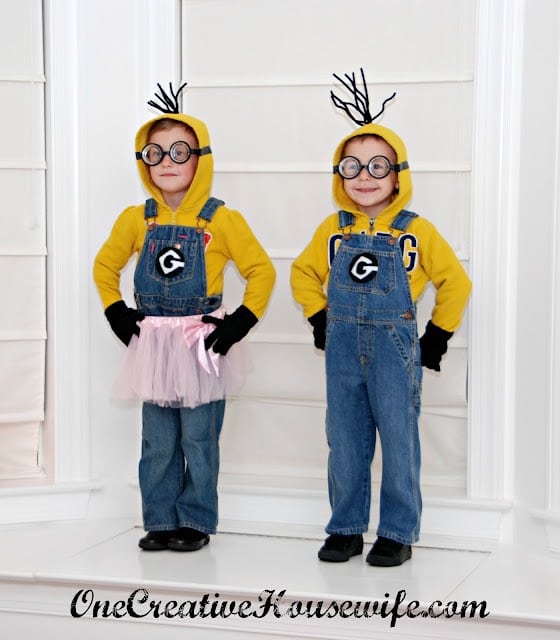 Two kids stand on a front porch wearing DIY Minion Halloween costumes consisting of blue denim overalls with simple black and white felt "Gru" symbols, black shoes and gloves, round black eyeglasses, and yellow hooded sweatshirts with black pipe cleaner hair coming out the top of the hoods. One also wears a pink tutu skirt over their overalls.