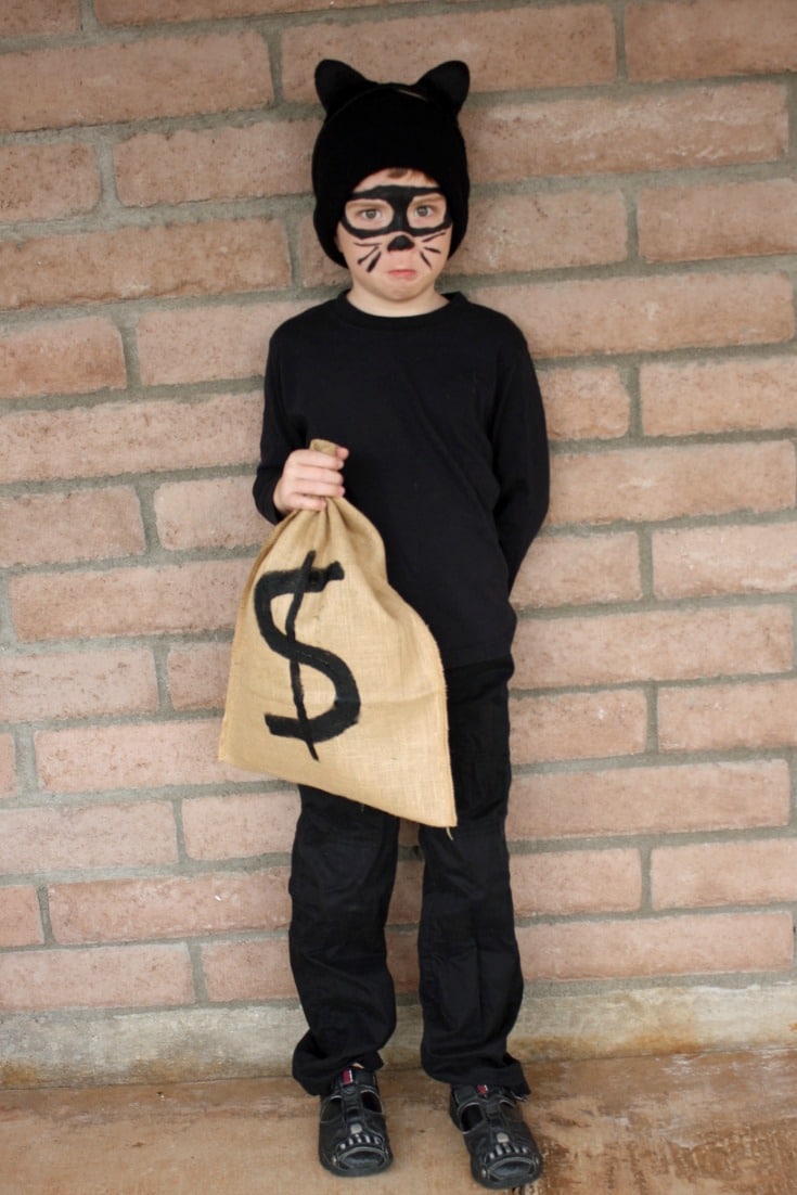 A child stands holding a burlap sack with a $ money symbol on it, wearing all black along with a black stocking cap with cat ears on it and black face makeup to look like whiskers, a cat nose, and an eye mask.