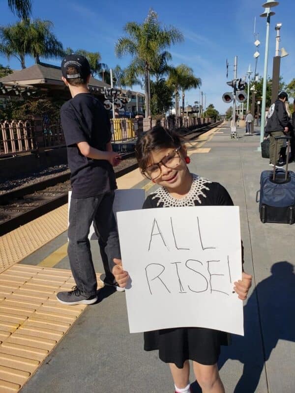 A photo of a smiling girl in a DIY Judge Ruth Bader Ginsburg costume consisting of a black dress, white crochet collar, big dark glasses, and holding a sign that says "All Rise!"