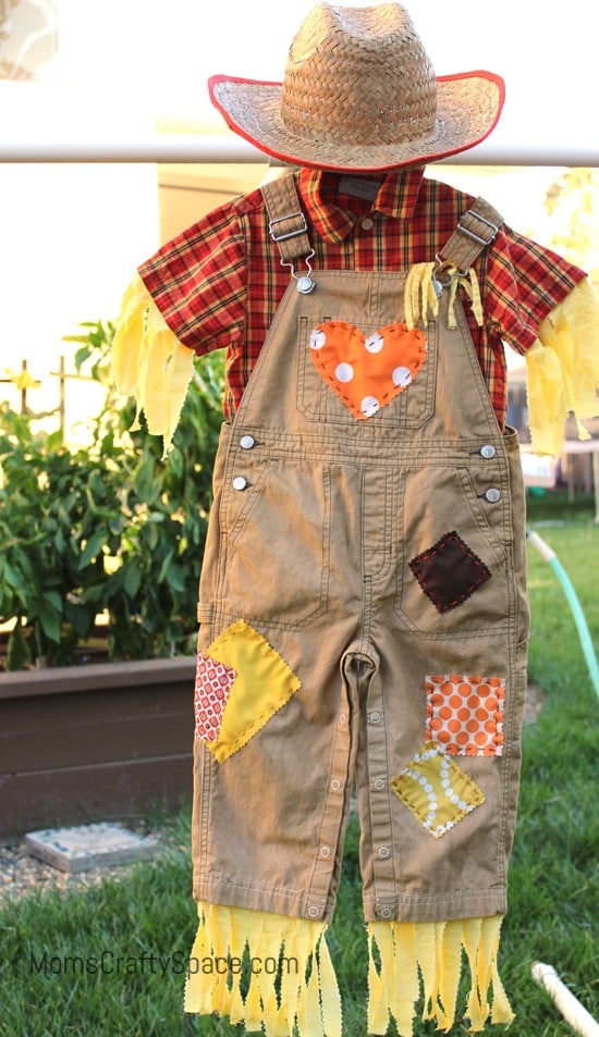 A homemade scarecrow Halloween costume hangs outside from a clothes hanger. The costume consists of tan overalls with multiple patterned fabric patches and a red plaid collar shirt with yellow fabric fringes coming out of the pant legs and sleeves along with a straw cowboy hat on top.