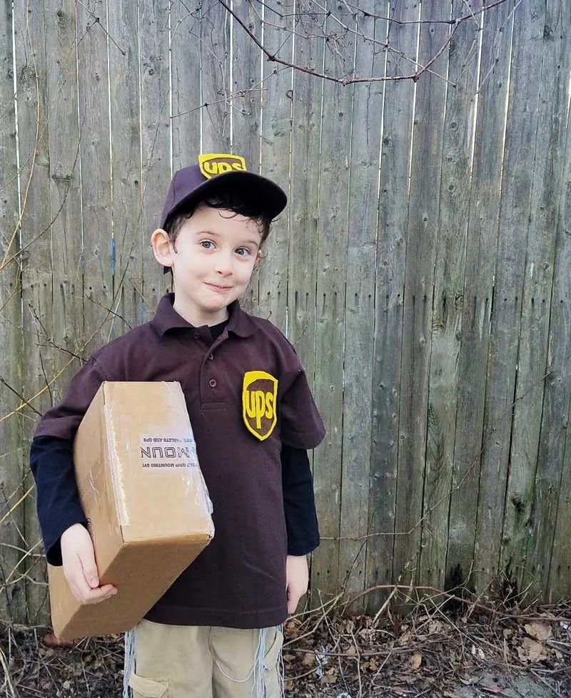 A young boy stands outside holding a cardboard box package and wearing a DIY UPS Driver Halloween costume consisting of tan khaki pants, a brown collared polo shirt with a DIY UPS logo attached to the chest, and a brown baseball cap with a DIY UPS logo attached to it.