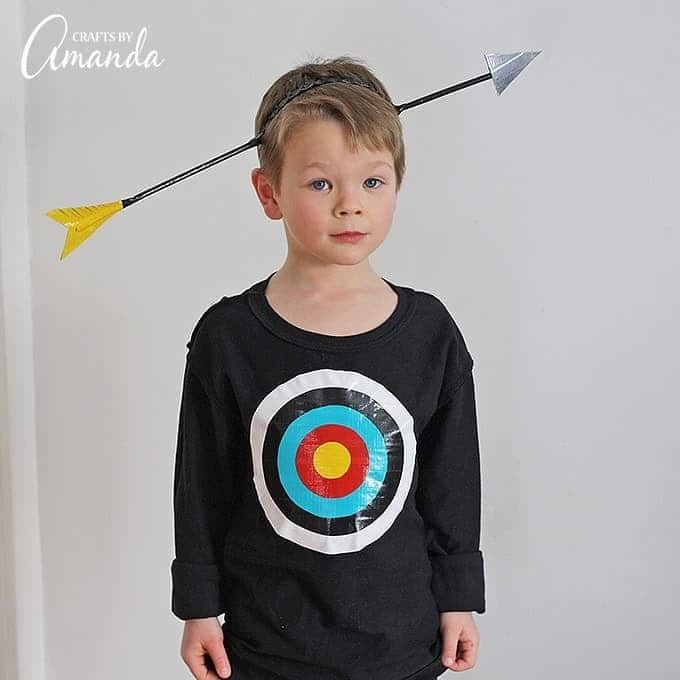 A child stands wearing a fun creative Halloween costume consisting of a black, long-sleeved t-shirt with a target in the center of it and a DIY headband that makes it look like an arrow is going through his head.