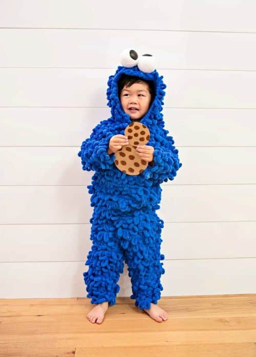 A smiling young child stands, wearing a creative DIY Cookie Monster Halloween costume made out of blue loop yarn, white plastic ball eyeballs, and cardboard cookies.