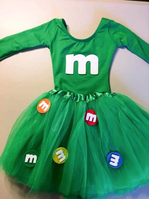 A DIY M&M Candy Halloween costume lays on a white surface. The costume consists of a long-sleeved green bodysuit with a white felt "m" in the center and a green tulle tutu with different colored felt M&M candies on it.