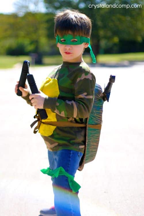 A young child stands outside wearing a DIY Ninja Turtle creative Halloween costume made out of denim blue jeans with green fabric strips tied around the legs, a green and tan camo long-sleeved t-shirt with yellow felt on the front and a painted disposable tin pan on the back for the turtle shell, a green felt eye mask, and pretend toy nunchucks.