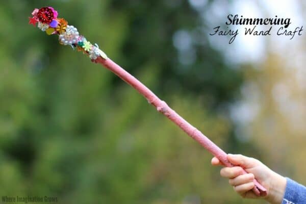 A photo of a child's hand holding a pink fairy wand crafted out of a stick, paint, and craft gems.