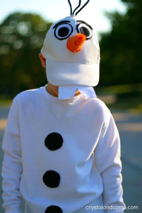 A child stands outside wearing a DIY Olaf Halloween costume consisting of a white sweatshirt with round black felt buttons down the center and a white baseball cap with an orange felt carrot nose, black and white eyes, and white felt teeth attached.