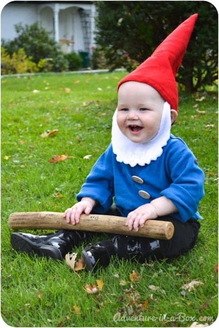 A smiling toddler sits in the grass wearing a homemade gnome Halloween costume including a pointed red felt hat, white felt beard, blue shirt, and black pants and boots.