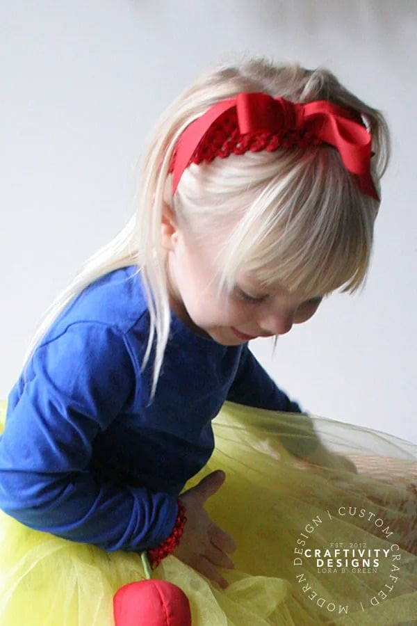 A photo of a smiling little girl with long blonde hair wearing a DIY Snow White costume made up of a homemade yellow tulle skirt, a long-sleeved royal blue shirt, and a big red hair bow.