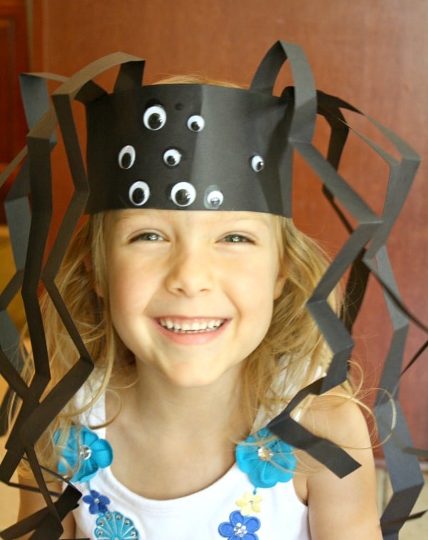 A photo of a smiling young girl wearing a spider headband craft for Halloween that she made out of black paper, accordion-folded black paper legs, and lots of googly eyes.