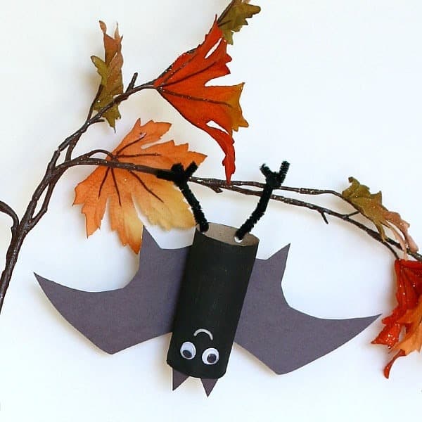 A photo of a hanging bat Halloween party craft for kids using a toilet paper tube, construction paper wings, and pipe cleaner legs.