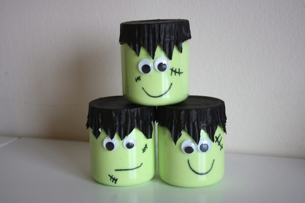 A photo of 3 DIY Franken-Slime Halloween party favors with light green slime inside clear plastic jars decorated with black cupcake liners cut into hair, googly eyes, and marker smiles and stitch marks.