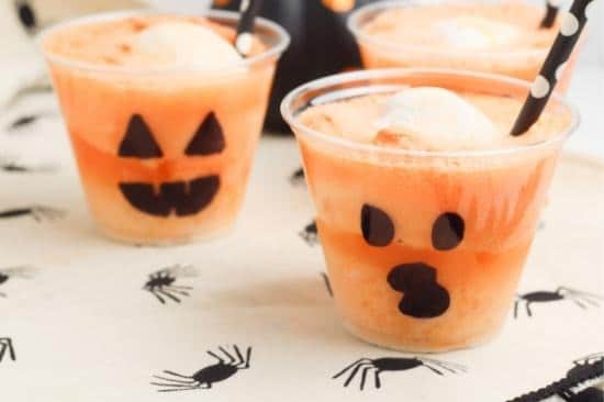 A photo of multiple Jack-O-Lantern Halloween Party Floats consisting of orange soda and vanilla ice cream inside clear plastic party cups decorated with black marker to look like Jack-O-Lanterns.