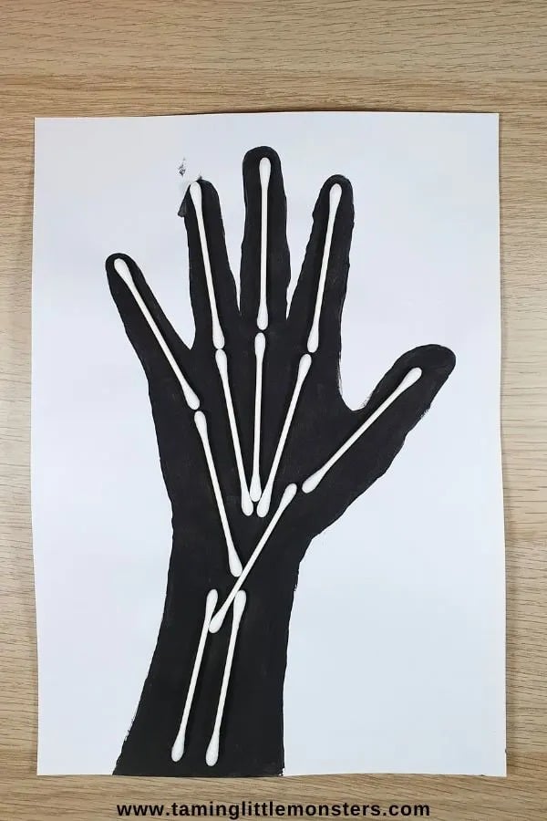 A photo of a kids skeleton handprint Halloween party craft featuring a black paper handprint on white paper with white cotton swabs glued onto it to look like bones.