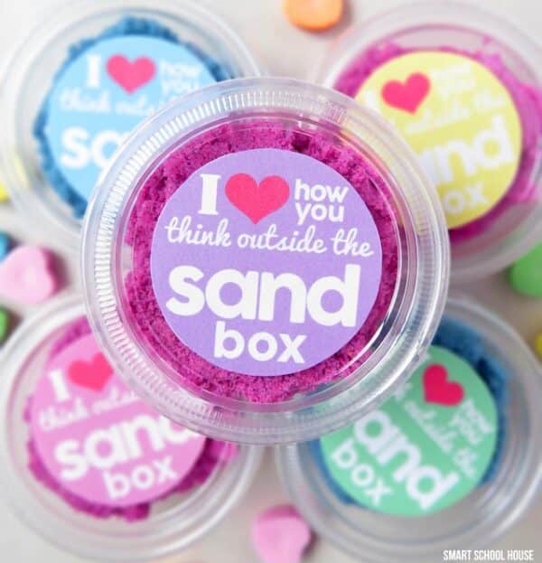 Fun DIY kinetic sand Valentines consisting of small plastic containers holding different colors of kinetic sand with the lids having printable labels saying "I love how you think outside the sandbox."