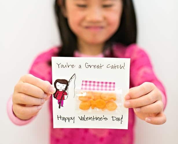 A happy girl holding up a cut printable Valentine featuring an illustration of a girl holding a fishing pole, the saying "You're a great catch!", and a small back of Goldfish Crackers taped onto it. A great non-candy Valentine idea for preschoolers.