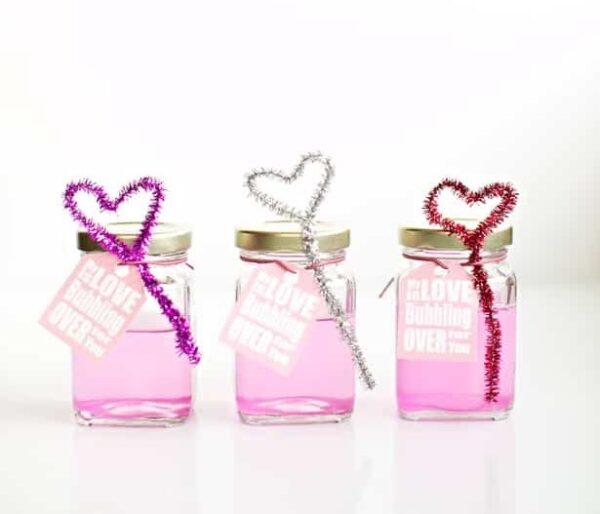 Three DIY pink bubbles and bubble wand Valentines consisting of mini glass mason jars holding homemade pink bubble solution with a heart-shaped bubble wand made out of pipe cleaner tied onto it. A great non-food Valentine idea for preschool kids.