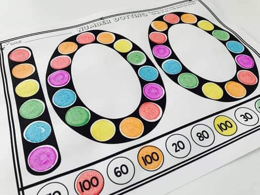 A photo of a printable 100th day of school math activity where students use dot markers to mark numbers that make 100.