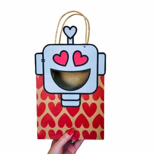 An easy paper bag Valentine mailbox idea featuring a hand holding up a red heart-patterned paper gift bag with a free printable robot graphic glued onto the top half with its smiling mouth cut out for Valentine cards to be put inside.