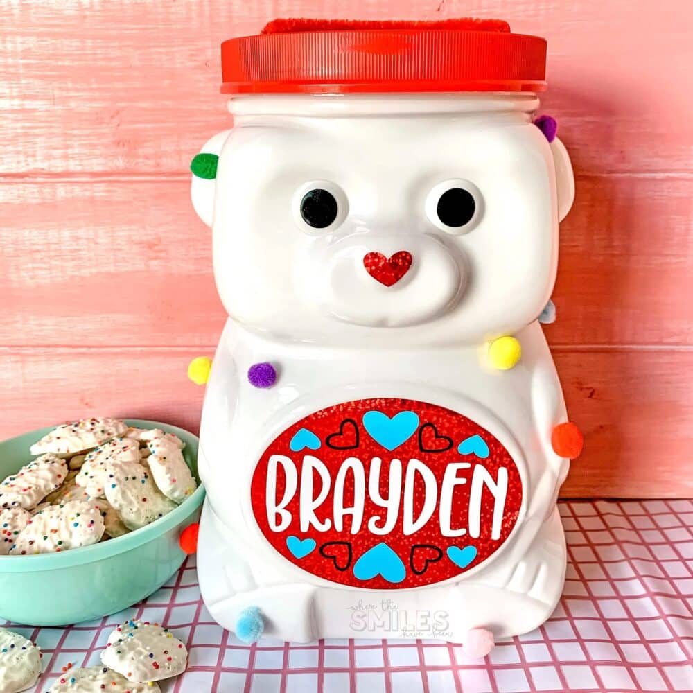 An easy bear Valentine box idea using a bear-shaped plastic animal cracker container painted white with colorful pom poms glued randomly around it, a red lid on top with a slip cut out of it, and vinyl decals applied for eyes, a heart nose, and a red label on the belly with blue hearts and the child's name, "Brayden."