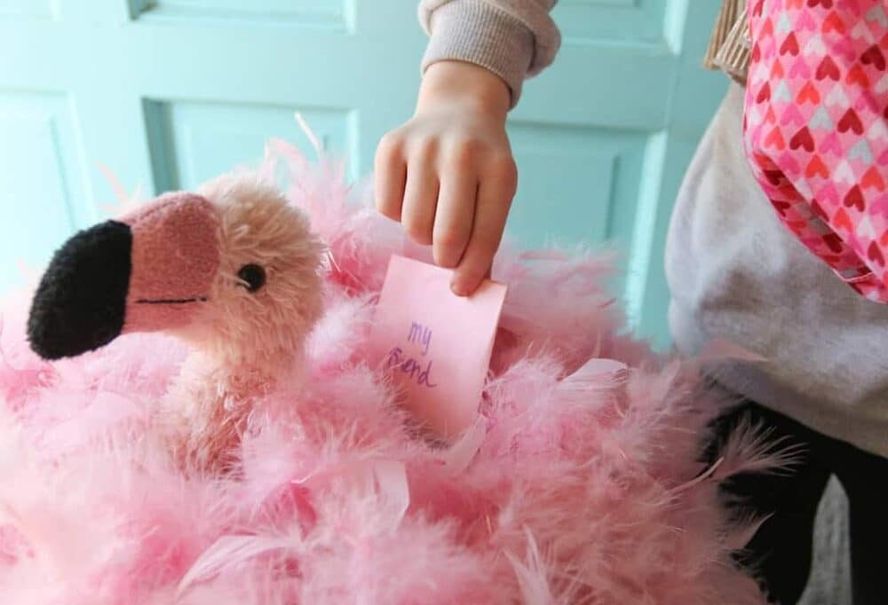 A cute DIY flamingo Valentine mailbox craft featuring a small child's hand putting a small pink paper Valentine card into a slot on the top of a homemade Valentine box made to look like a pink flamingo using a plush flamingo toy head and pink feather boas.