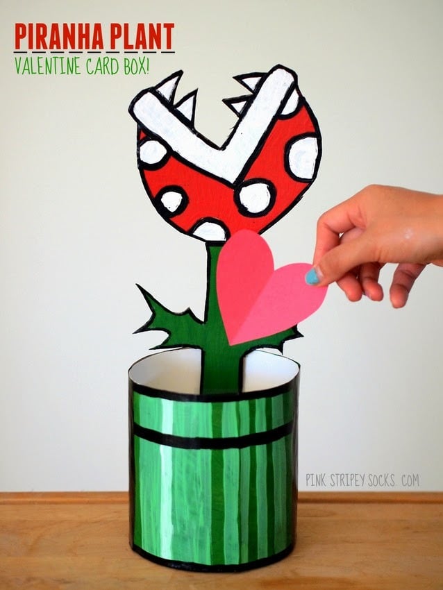 A cool Nintendo Piranha Plant Valentine Card Box idea for kids featuring a hand putting a heart-shaped paper Valentine card into the opening of a Valentine box made to look like a red, white, and green chomping piranha plant from a Nintendo video game using paint, cardboard, paper, and an oatmeal canister.