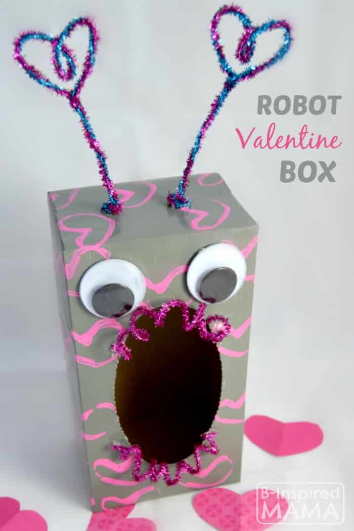 A cool robot Valentine box idea featuring a tissue box standing on its end with opening facing the viewer, painted gray with pink stamped heart shapes, with big googly eyes above the opening, twirly sparkly pipe cleaners on the top and bottom of the opening, and two tall sparkly pipe cleaner antennae coming out of the top with heart shapes on the ends.