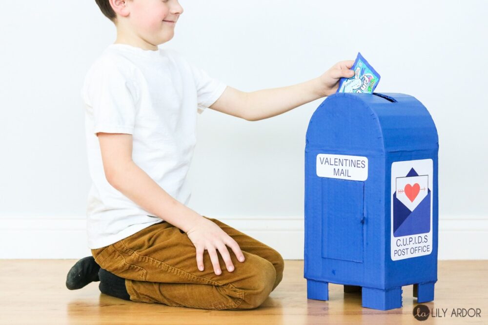 A creative DIY Valentine Mailbox idea using cardboard constructed to look like a post office mailbox, painted blue, with printable labels saying "Valentine Mail" and "CUPIDS Post Office." A happy child sits on his knees to the left of the box and extends an arm to put a small paper Valentine card into the slot on the top of the post office box.